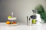 Sana 727 Supreme cold press juicer in Soft White with a glass of green juice and a fruit stand